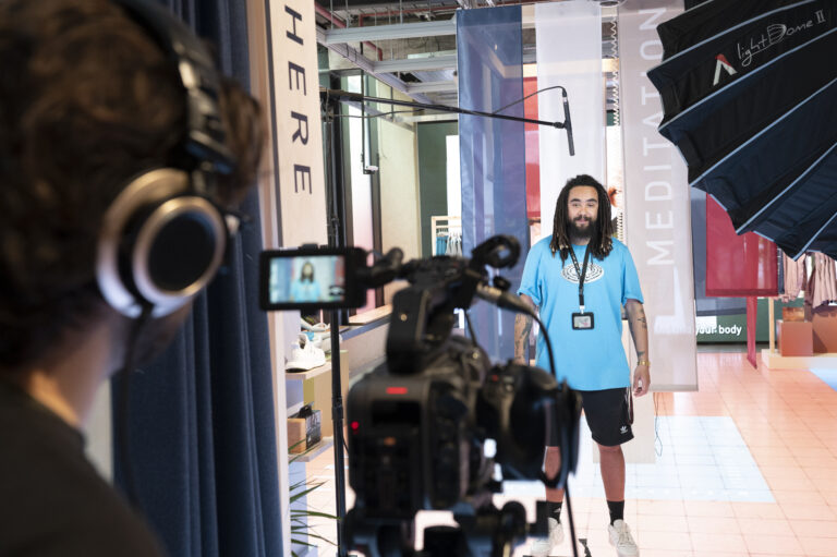 Viewed from behind the videographer. The videographer is filming a member of Adidas staff. The videographer is wearing headphones and looking into the view finder of the camera. You can also see other video equipment, including a light umbrella.