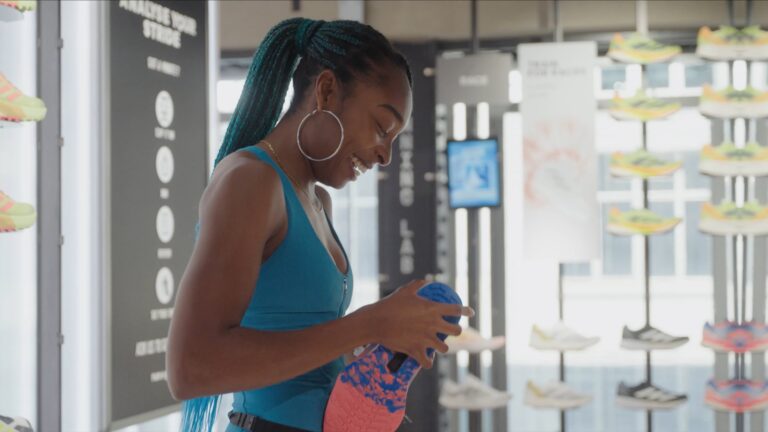 A member of Adidas staff in an Adidas store holding a brightly coloured trainer. There are columns of blurred out trainers in the background.