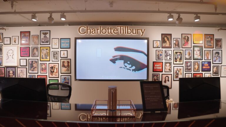A screen grab from a case study video showing a wall in the Charlotte Tilbury offices. There's a big screen in the centre and a big gallery wall of magazine covers, quotes prints and photos of the faces of women.
