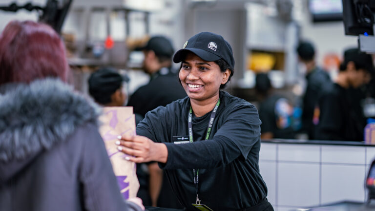 A member of staff from Taco Bell handing over a bag of food to a customer. She is smiling at the customer and wearing a Welcome Break lanyard.
