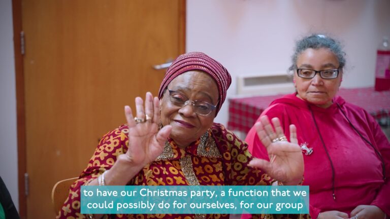 A screen grab from a video showing two older ladies. One lady is wearing a scarf on her head and a vibrant shirt with sparkly collar points. She has both her hands up mid-wave and is looking at the camera smiling. The other lady is sitting next to her and looking at her.